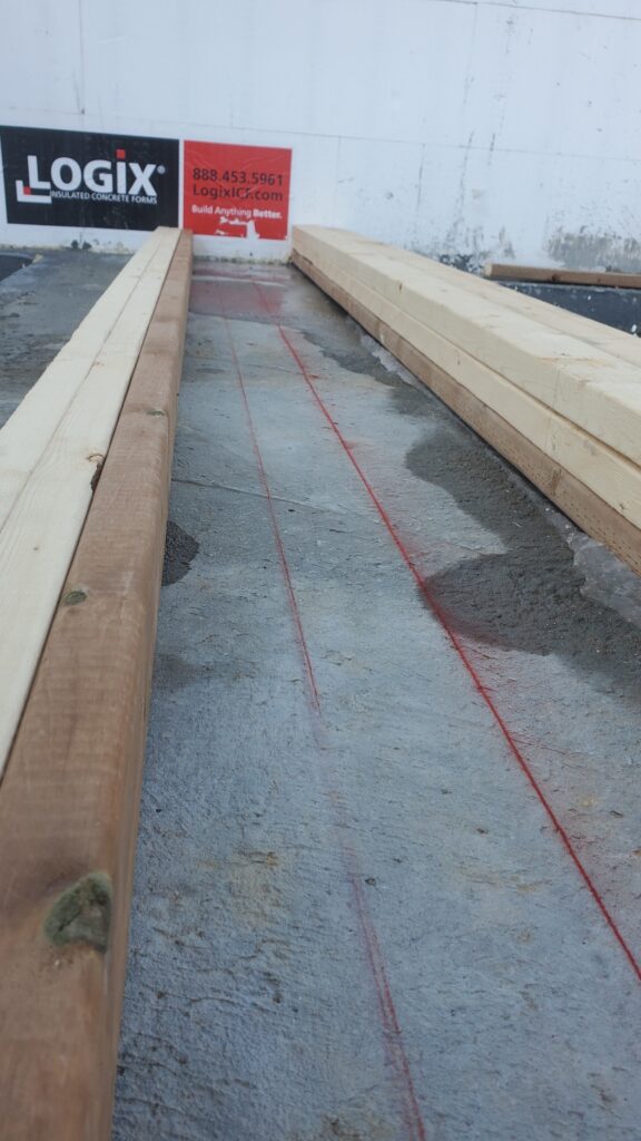 Red chalk lines marking on a gray concrete footing for precise wood load-bearing installation.