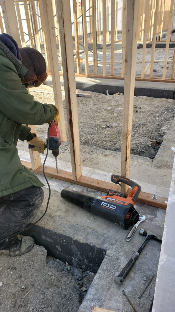 Laborer drilling a hole through the bottom plate of a party wall during strip footing installation, showcasing precision craftsmanship for a solid foundation.